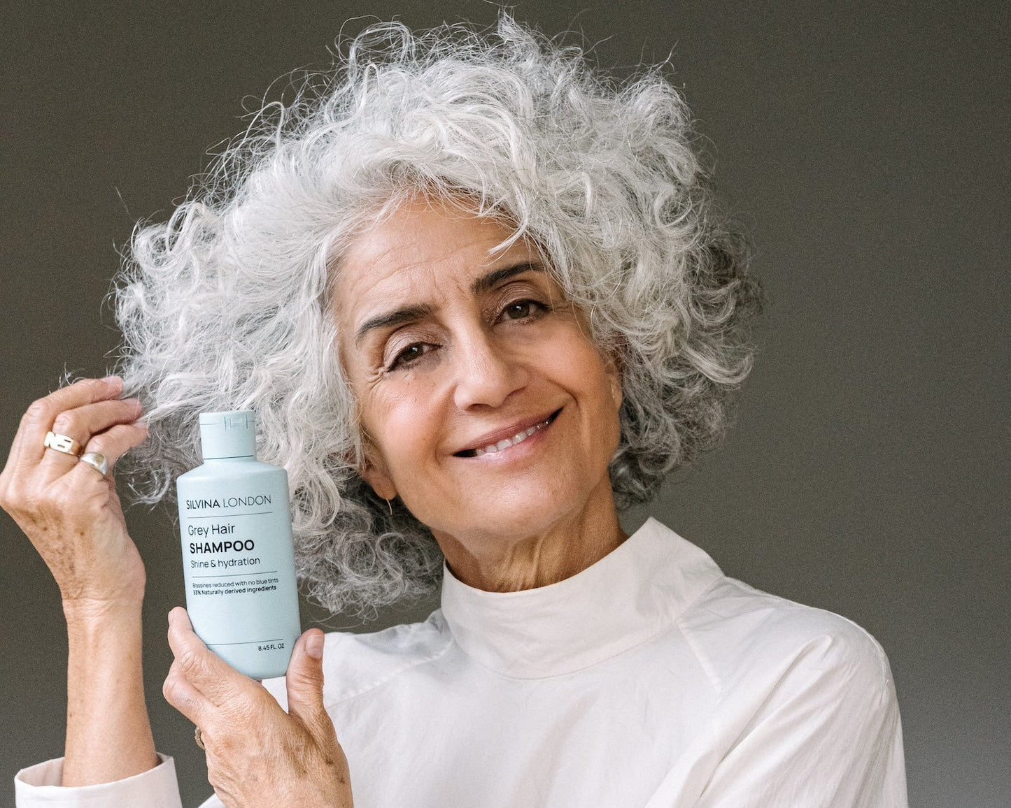 Woman with grey hair holding a bottle of Grey Hair Shampoo by Silvina London against a neutral background.