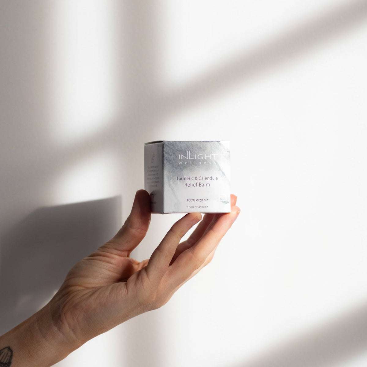 Hand holding a small container labeled Inlight Beauty Arnica & Hyssop Trauma Balm 45ml against a neutral background