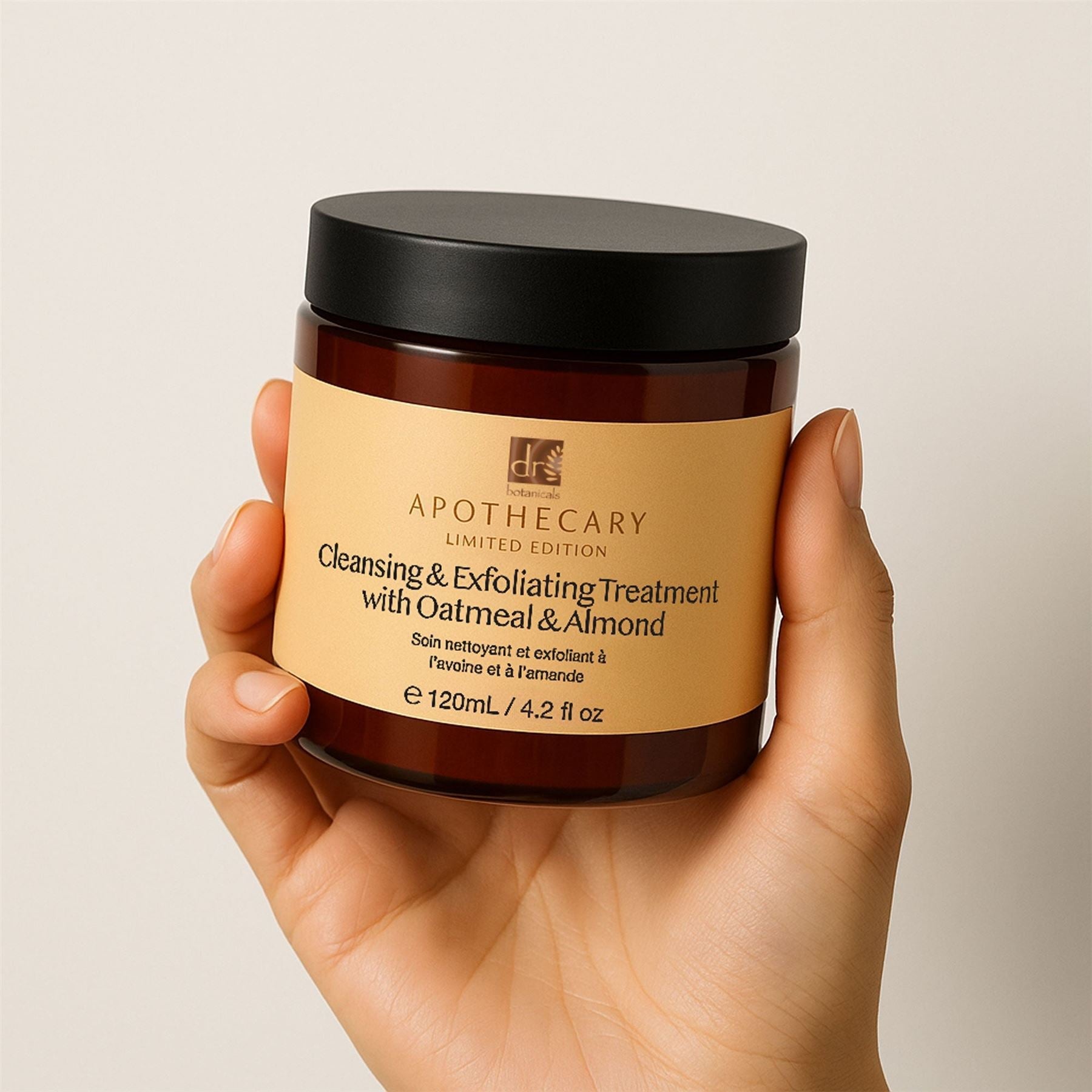 Hand holding a jar of 'Apothecary' cleansing and exfoliating treatment with oatmeal and almond on a beige background