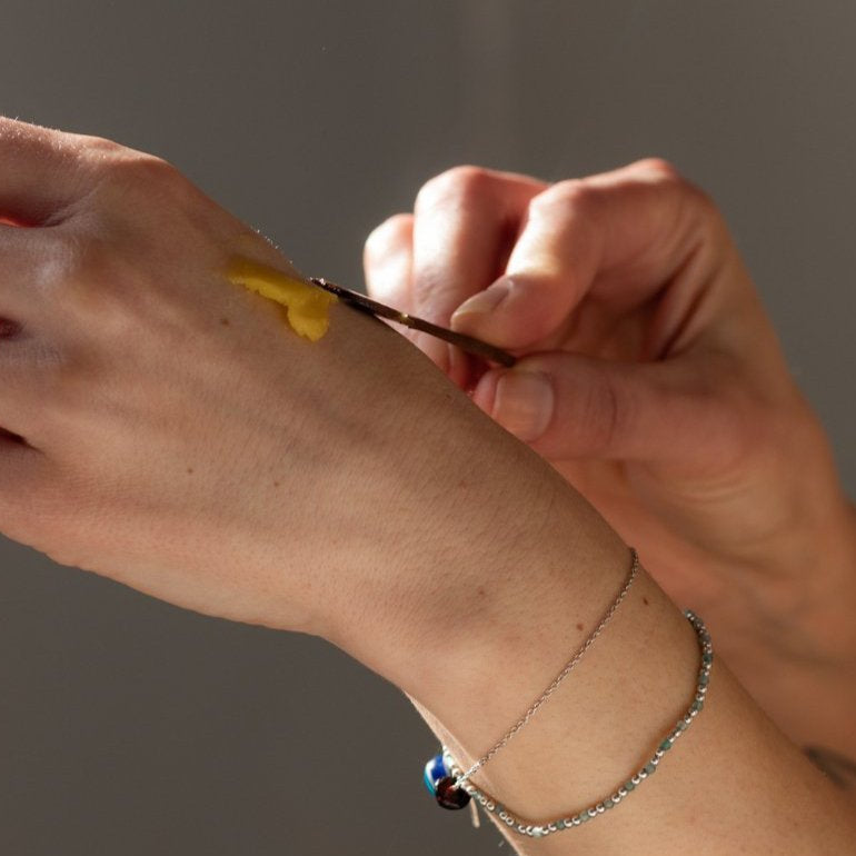 Close-up of hands applying Inlight Beauty Turmeric & Calendula Relief Balm 45ml to a surface with a brush on a grey background