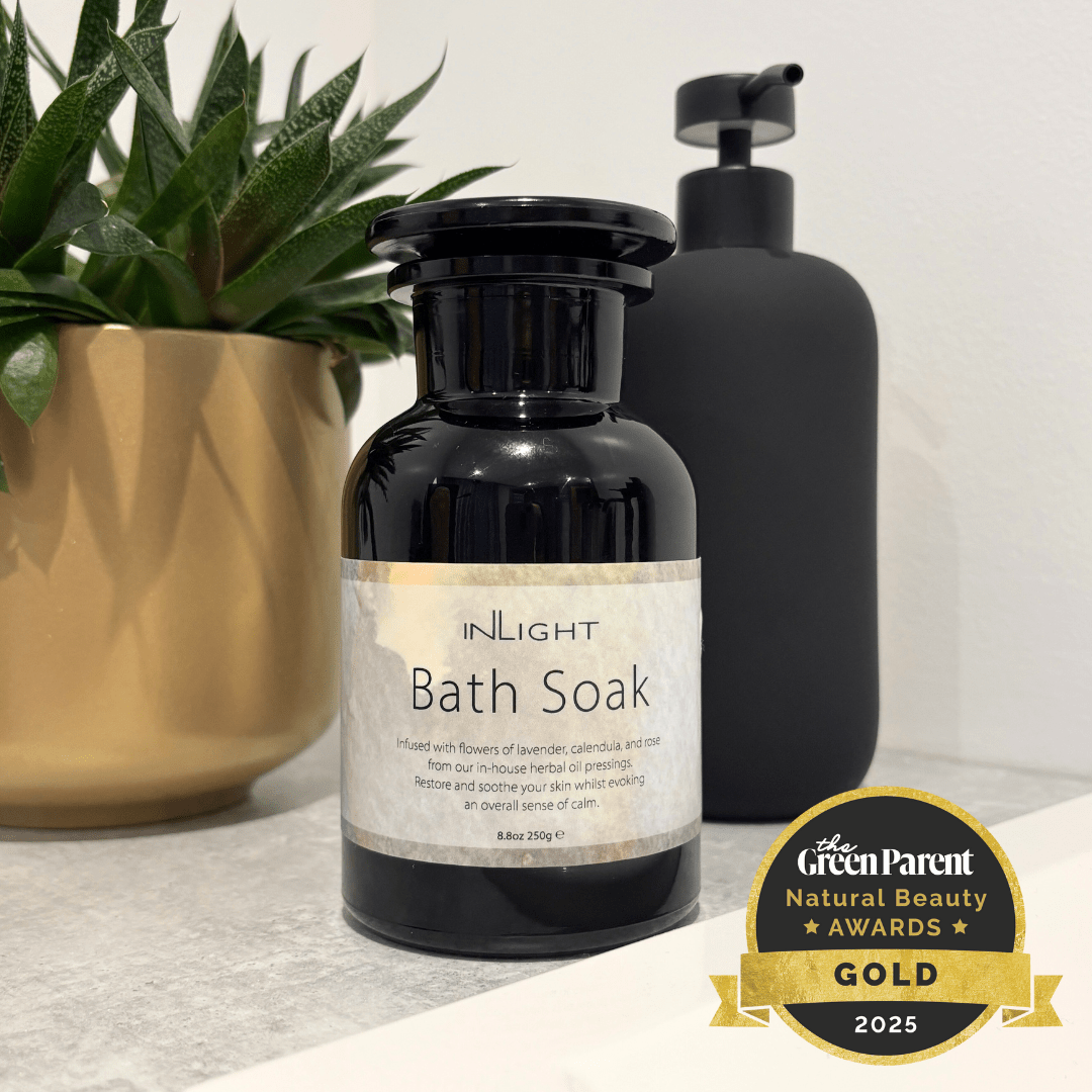 InLight Bath Soak in a black glass bottle with a cream label, styled on a marble countertop next to a potted plant and a matte black pump bottle, with a Gold 2025 award badge in the corner.