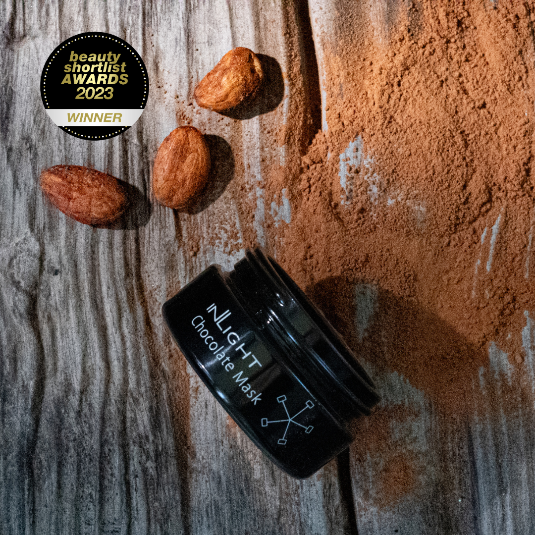 A black jar of Inlight Beauty Chocolate Mask 25ml lies on a rustic wooden surface with cocoa powder and three almonds scattered nearby. A circular 'beauty shortlist awards 2023 winner' badge appears in the upper left.