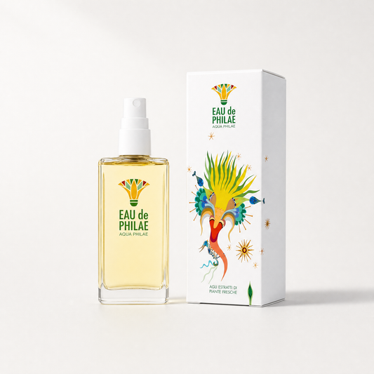 A clear glass spray bottle filled with pale yellow liquid stands beside a tall white product box. Both display the brand name EAU de PHILAE and a colourful illustrated crest with green text reading AGUA PHILAE.