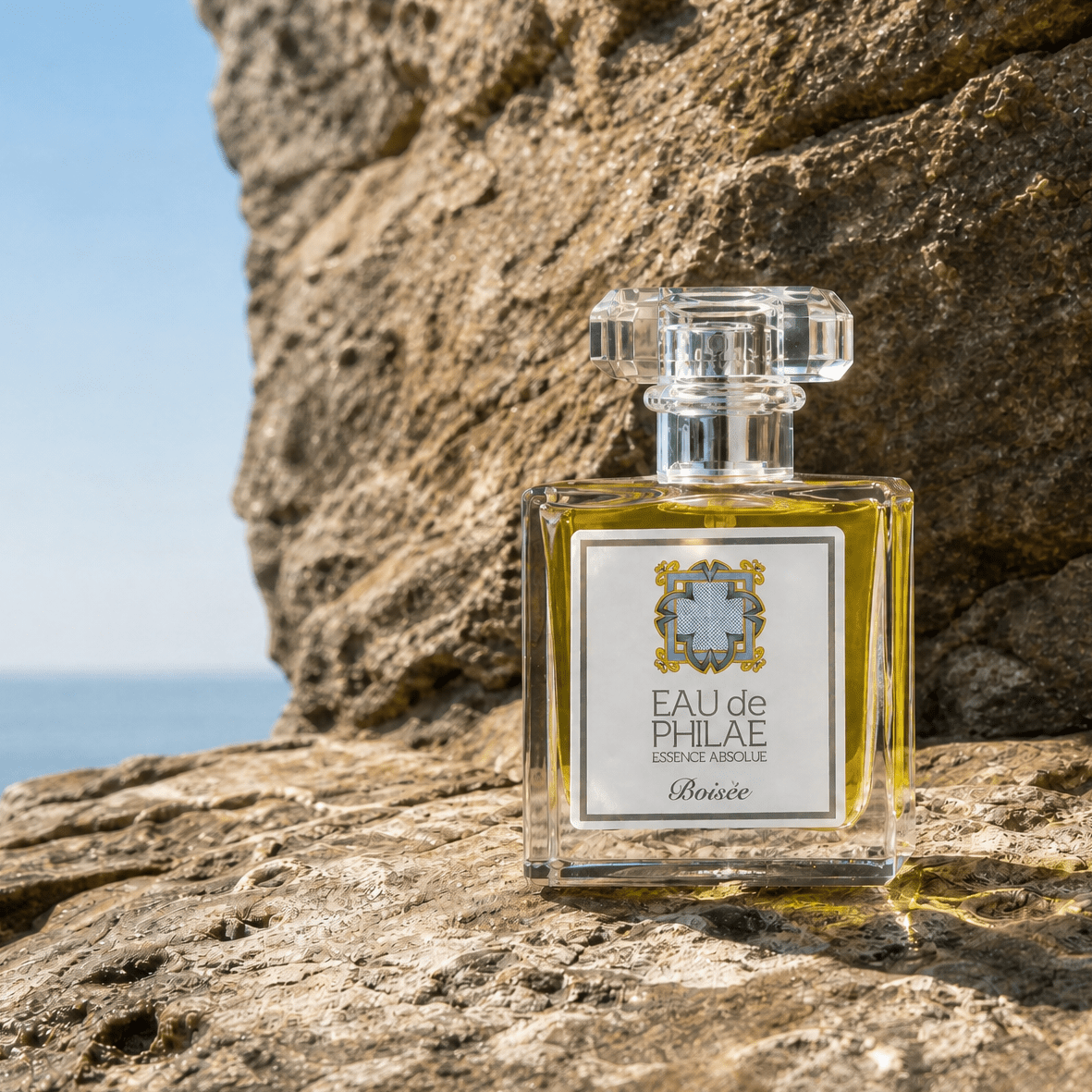 A clear glass perfume bottle with pale yellow liquid and a white label reading 'EAU de PHILAE' and 'Rosee', placed on a rocky beach with the sea and sky in the background.