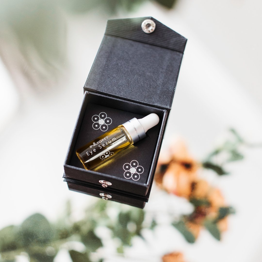 A small amber dropper bottle labeled 'EYE SERUM' resting inside an open black presentation box with floral printed details, photographed against a softly blurred light background with greenery and flowers.