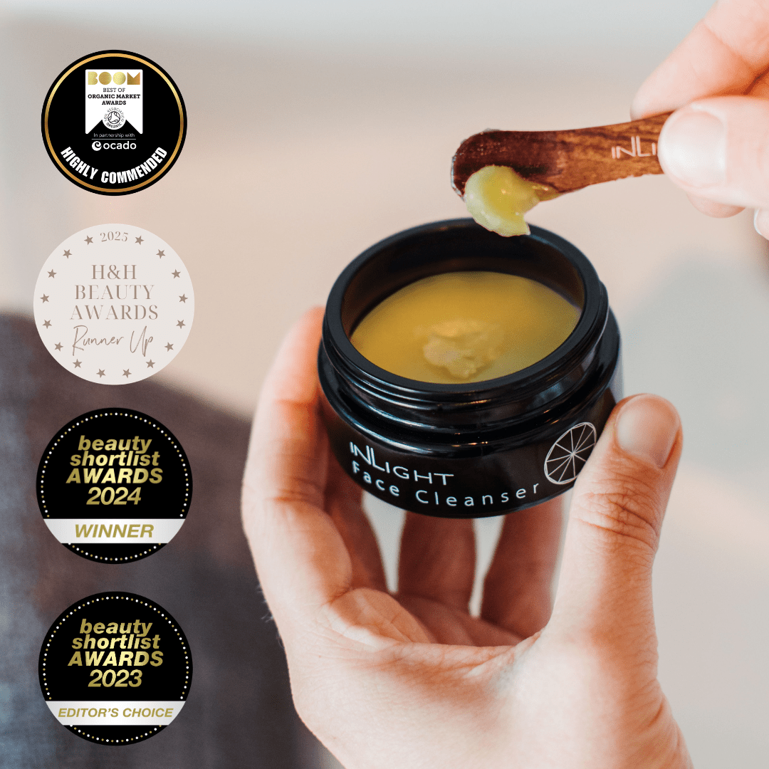 Hand holding a black jar of face cleanser with a wooden spoon, surrounded by awards on a blurred background.