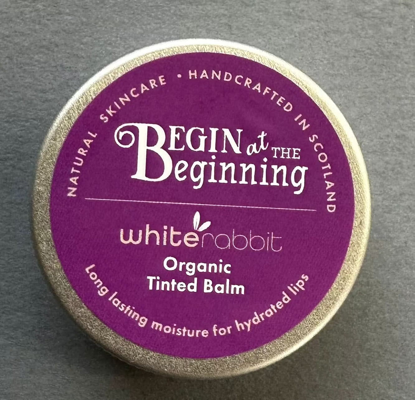 Lip balm container with 'Begin at the Beginning' branding on a grey background