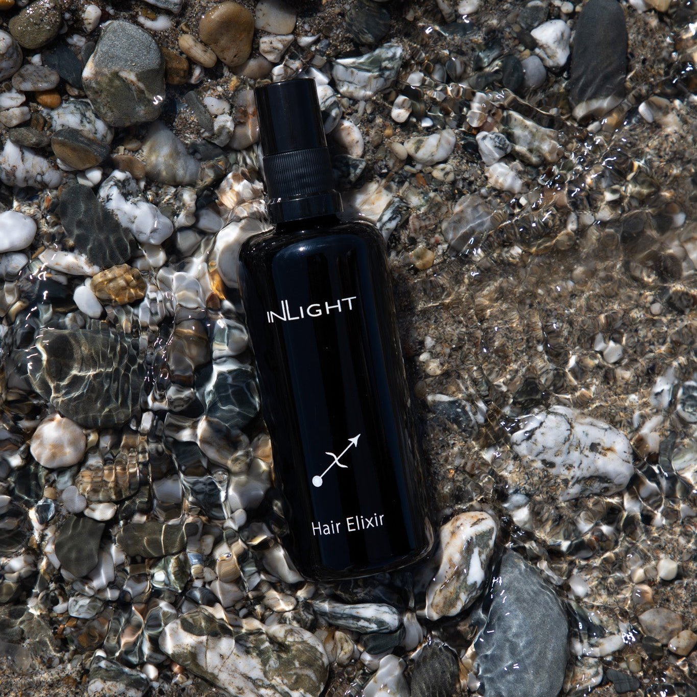 Black bottle labeled Inlight Beauty Hair Elixir 100ml on a pebbly surface