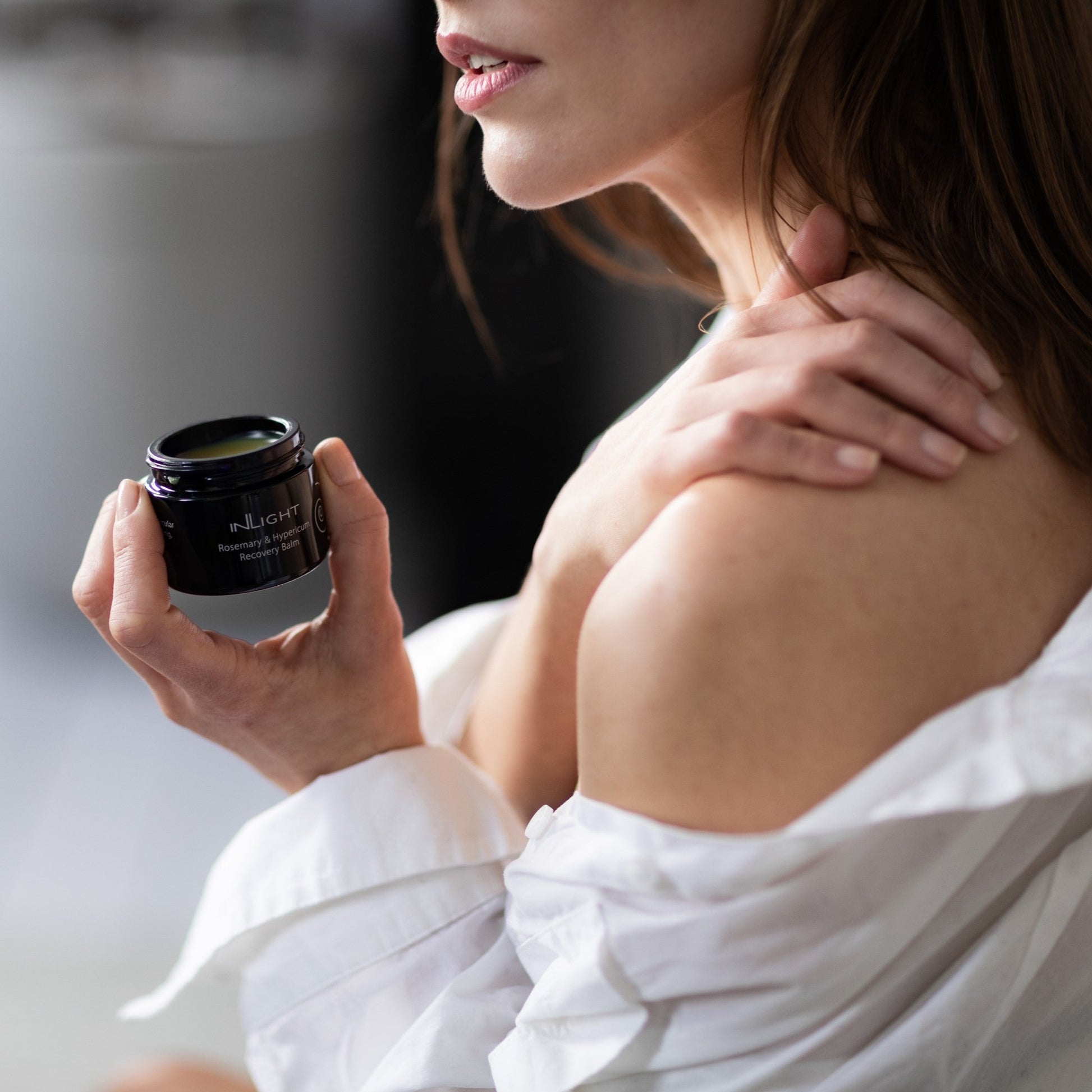 Woman applying Inlight Beauty Rosemary & Hypericum Recovery Balm 45ml to her shoulder with a jar of cream in her hand, wearing a white robe.