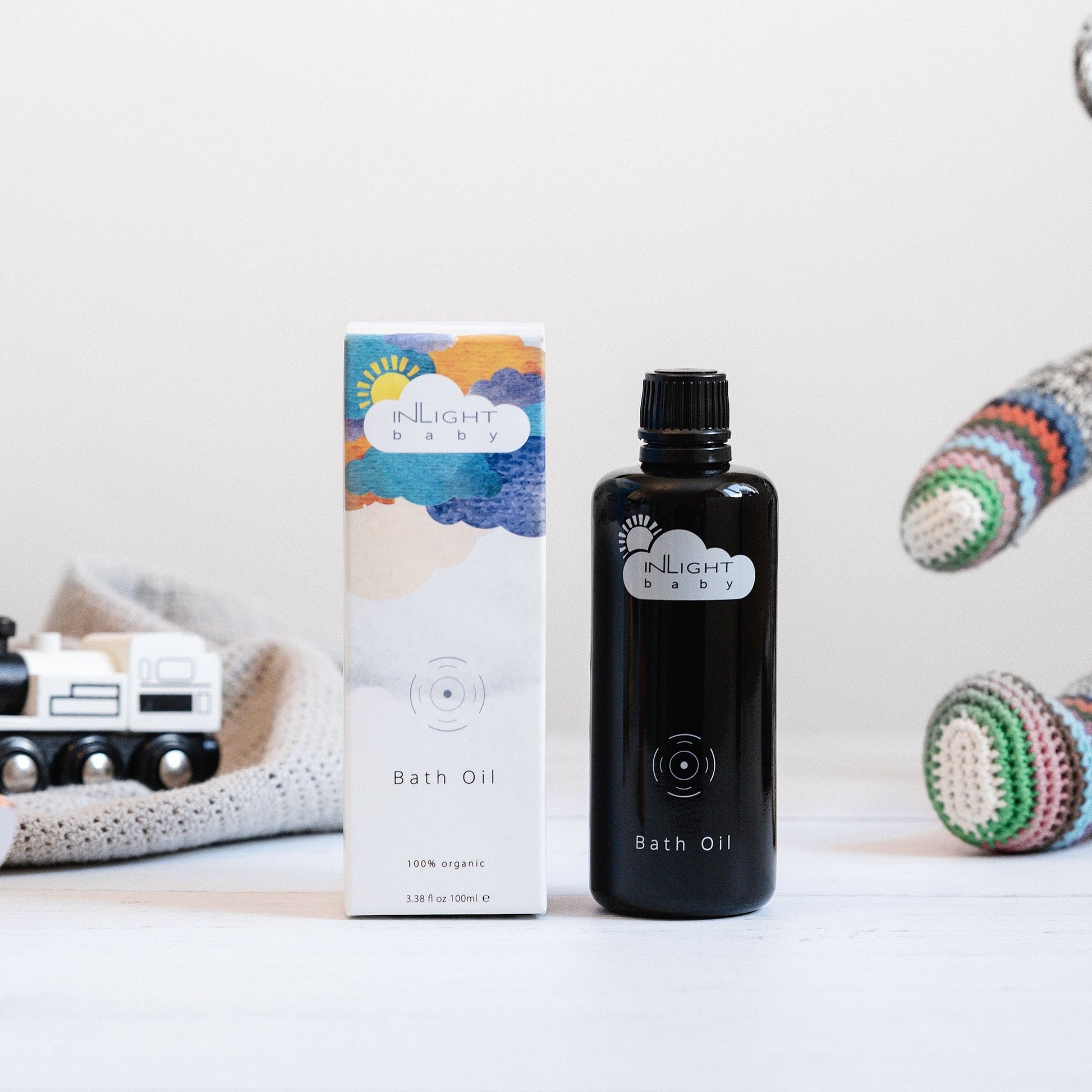Black bottle and colourful box of Inlight Beauty Baby Bath Oil 100ml on a white surface with toys in the background.