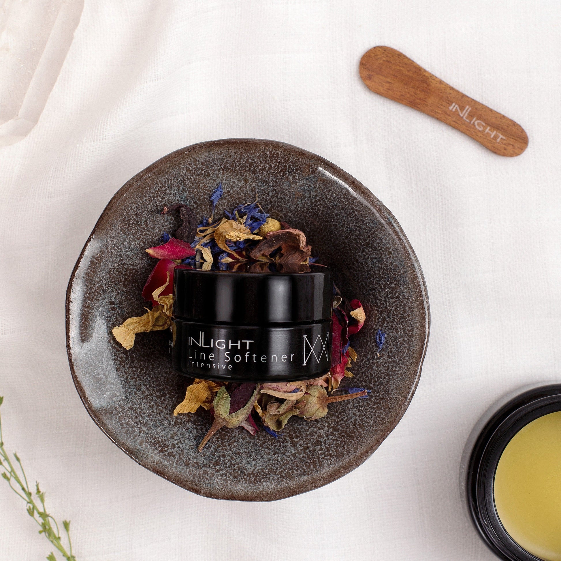 Black jar labeled 'Inlight Beauty Line Softener Intensive 15ml' on a textured plate with dried flowers, on a white background.