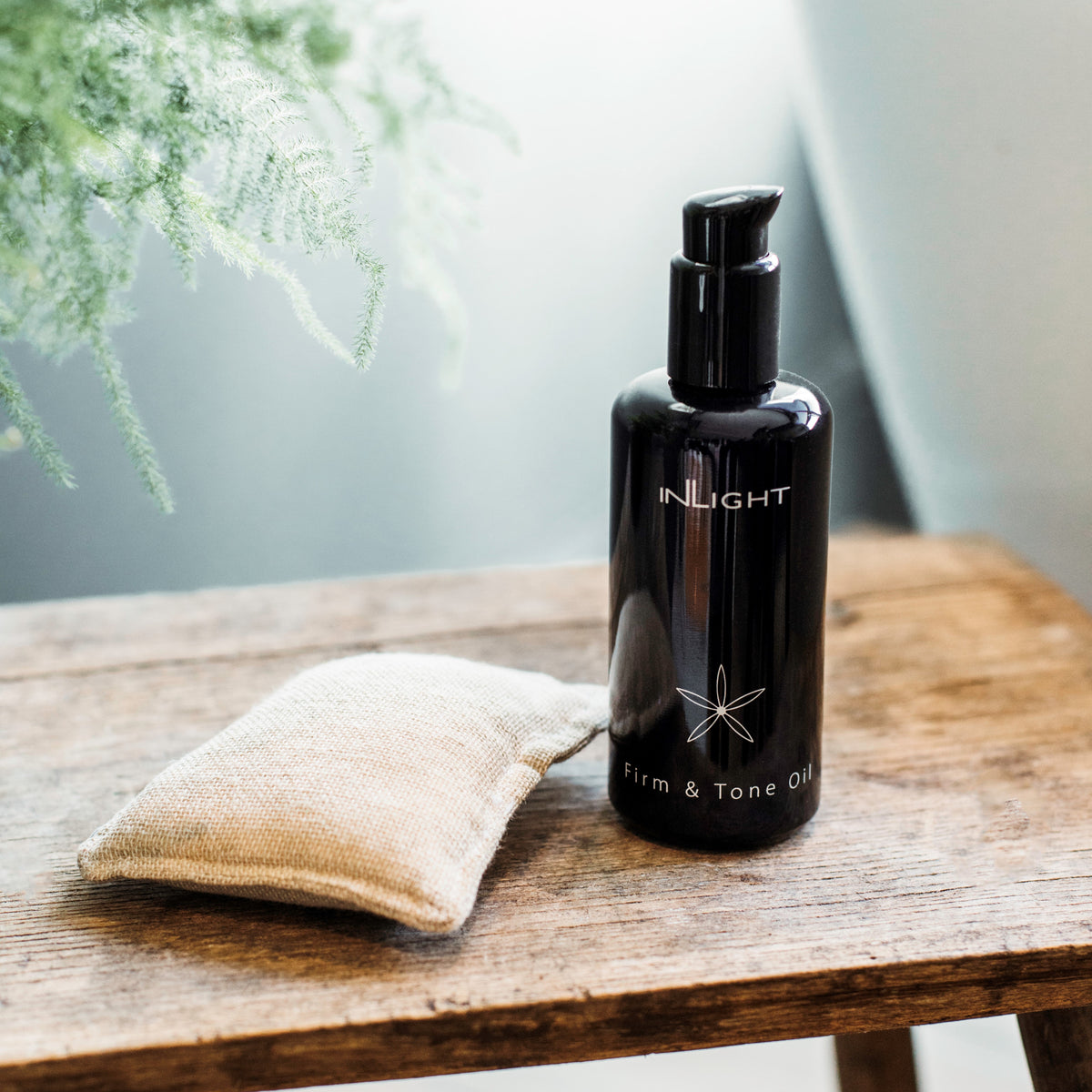 Black bottle of Inlight Beauty Firm & Tone Oil 200ml on a wooden surface with a textured white cloth and blurred greenery in the background.