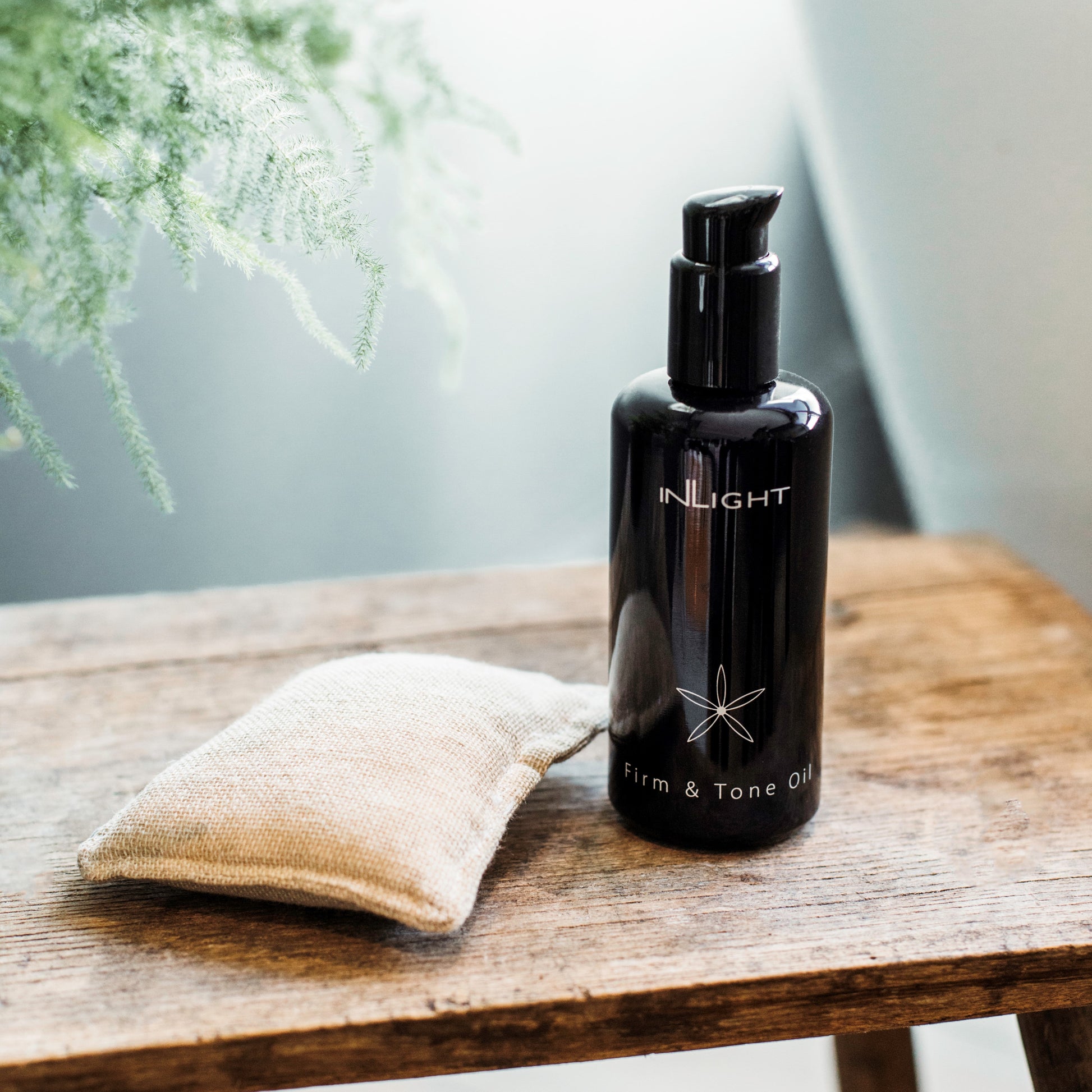 Black bottle of Inlight Beauty Firm & Tone Oil 200ml on a wooden surface with a textured white cloth and blurred greenery in the background.