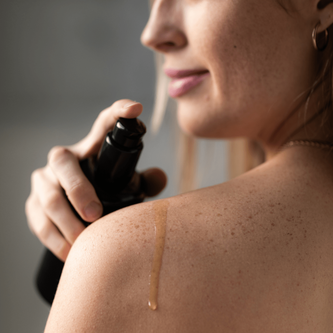 Person applying Inlight Beauty Body Oil with Arnica to shoulder with a black bottle against a neutral background