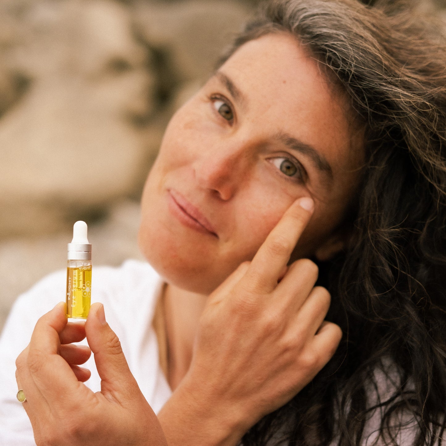 A woman holding a small dropper bottle of Inlight Beauty Eye Serum 4.9ml, touching her cheek as if applying the product, with a soft-focus outdoor background.
