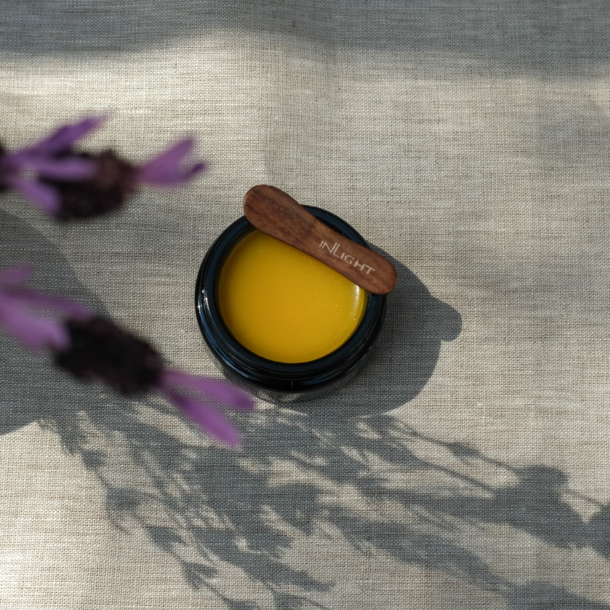 Jar of Inlight Beauty Night Balm 45ml with a wooden applicator on a textured surface with purple flowers.