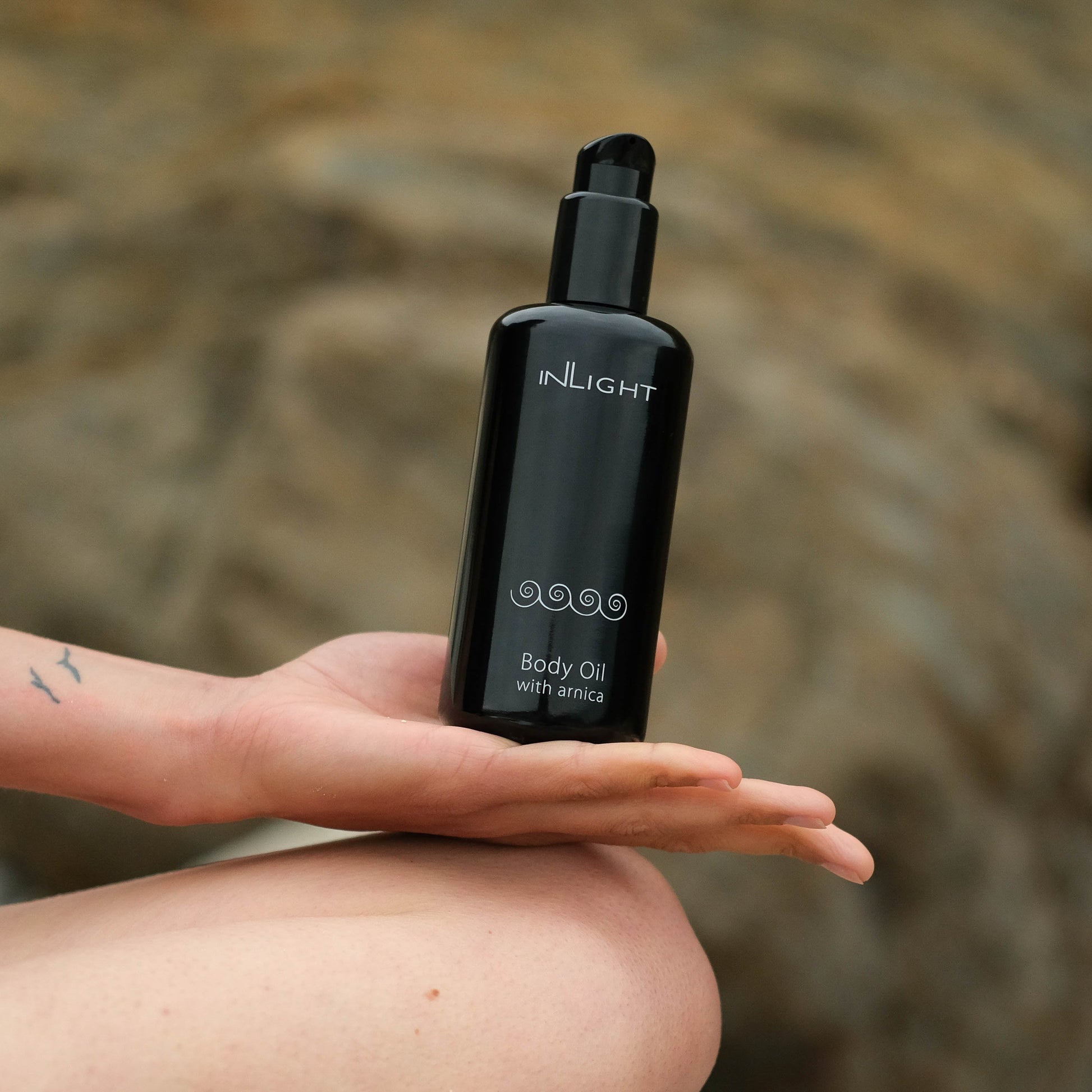 A hand holds a black pump bottle of inLIGHT Body Oil with arnica against a blurred natural rocky background.