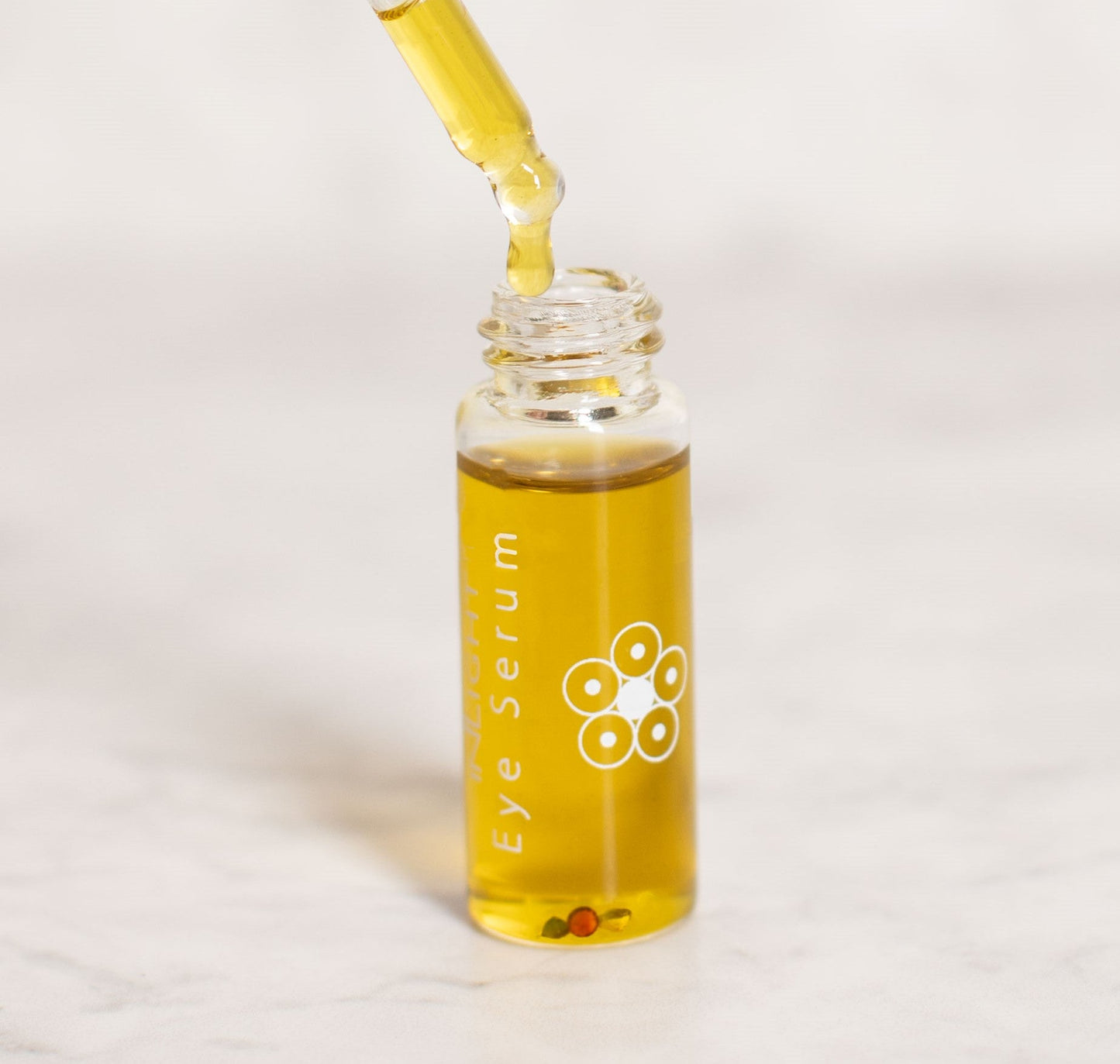 A small clear glass bottle of Inlight Beauty Eye Serum 4.9ml with golden yellow liquid, shown with a glass dropper dispensing more serum onto a light marble surface.