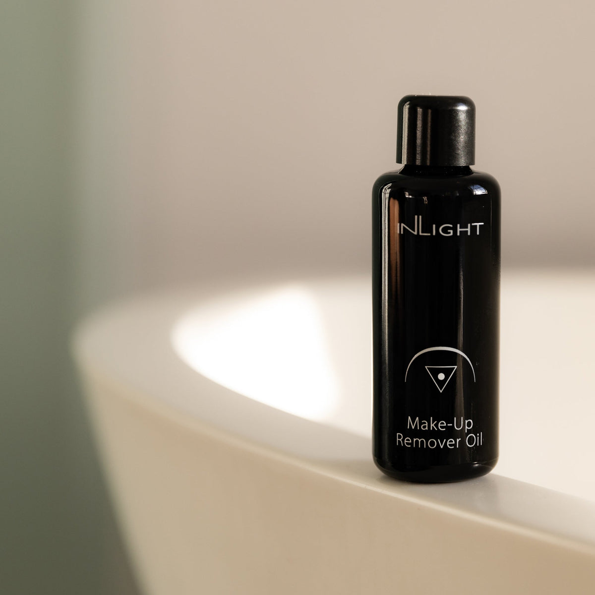 A black bottle of Inlight Beauty Make-up Remover Oil 50ml standing on the rim of a white sink in a softly lit bathroom setting.