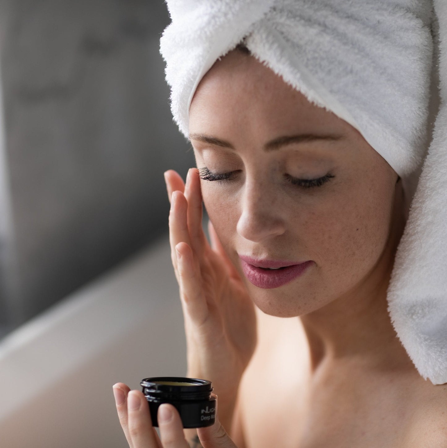 A woman with a white towel wrapped around her hair applies Inlight Beauty Deep Moisture Balm 28ml from a small black jar to her face in a bathroom setting.