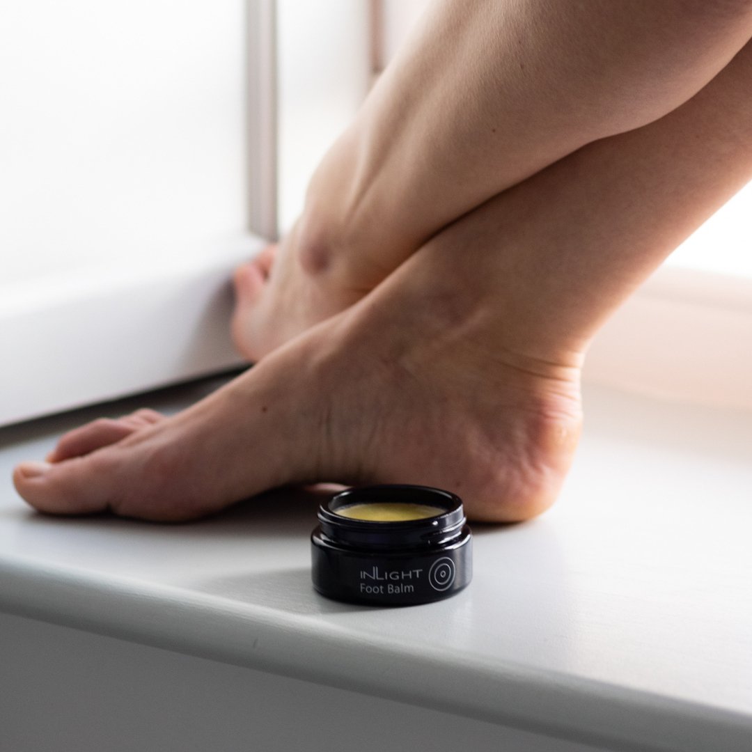 Person applying foot balm to their feet with a jar of Inlight Beauty Foot Balm 45ml on a white surface.