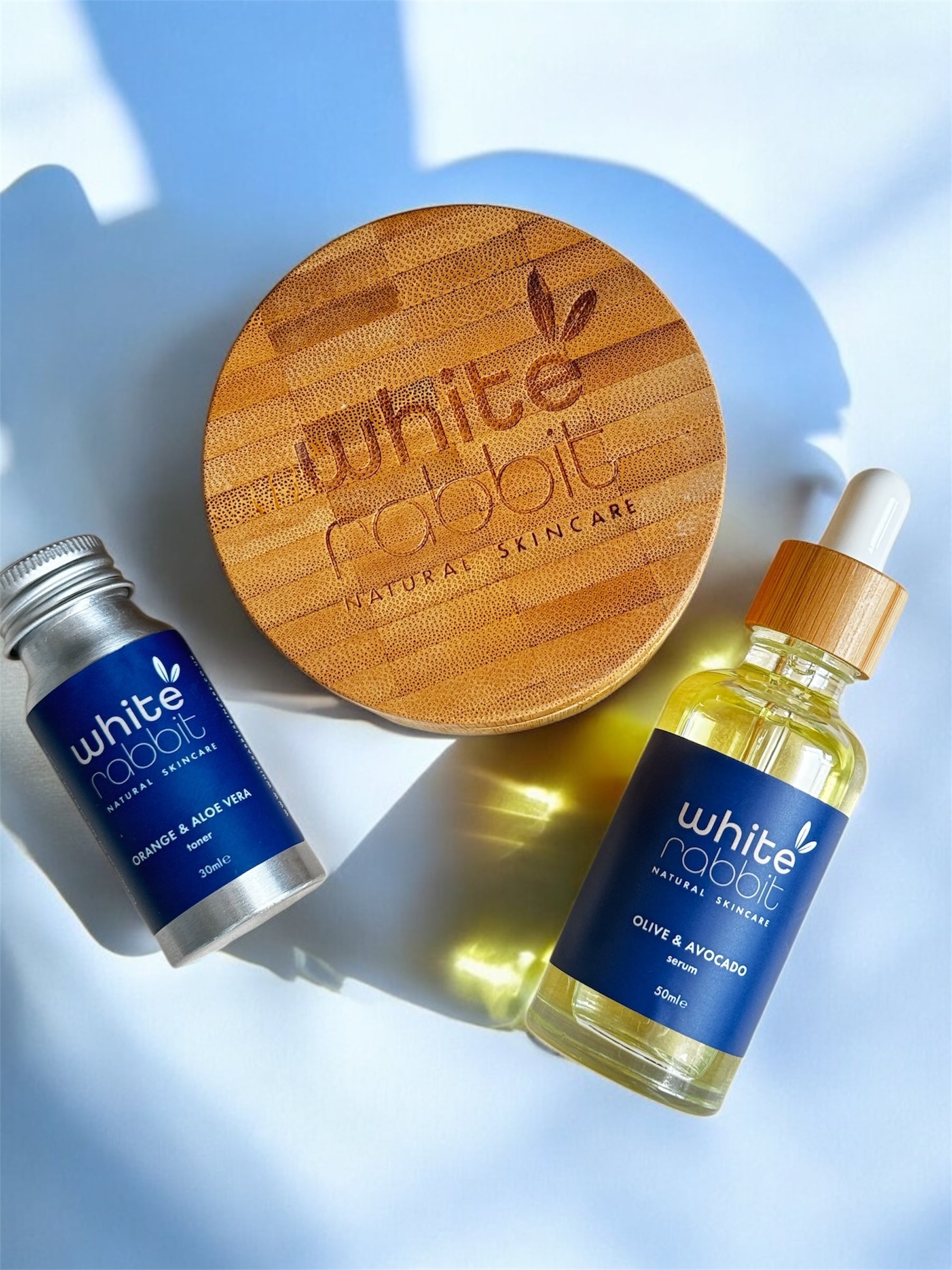 White Rabbit skincare products including a wooden disc, small tube, and bottle on a light blue background.