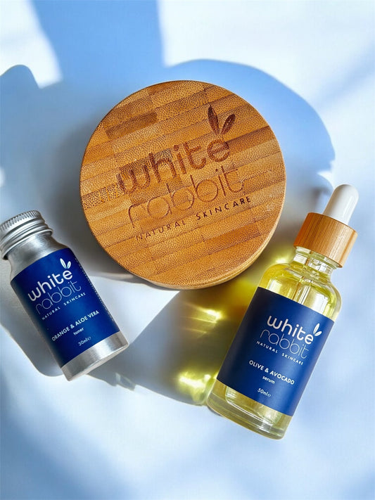 White Rabbit skincare products including a wooden disc, small tube, and bottle on a light blue background.