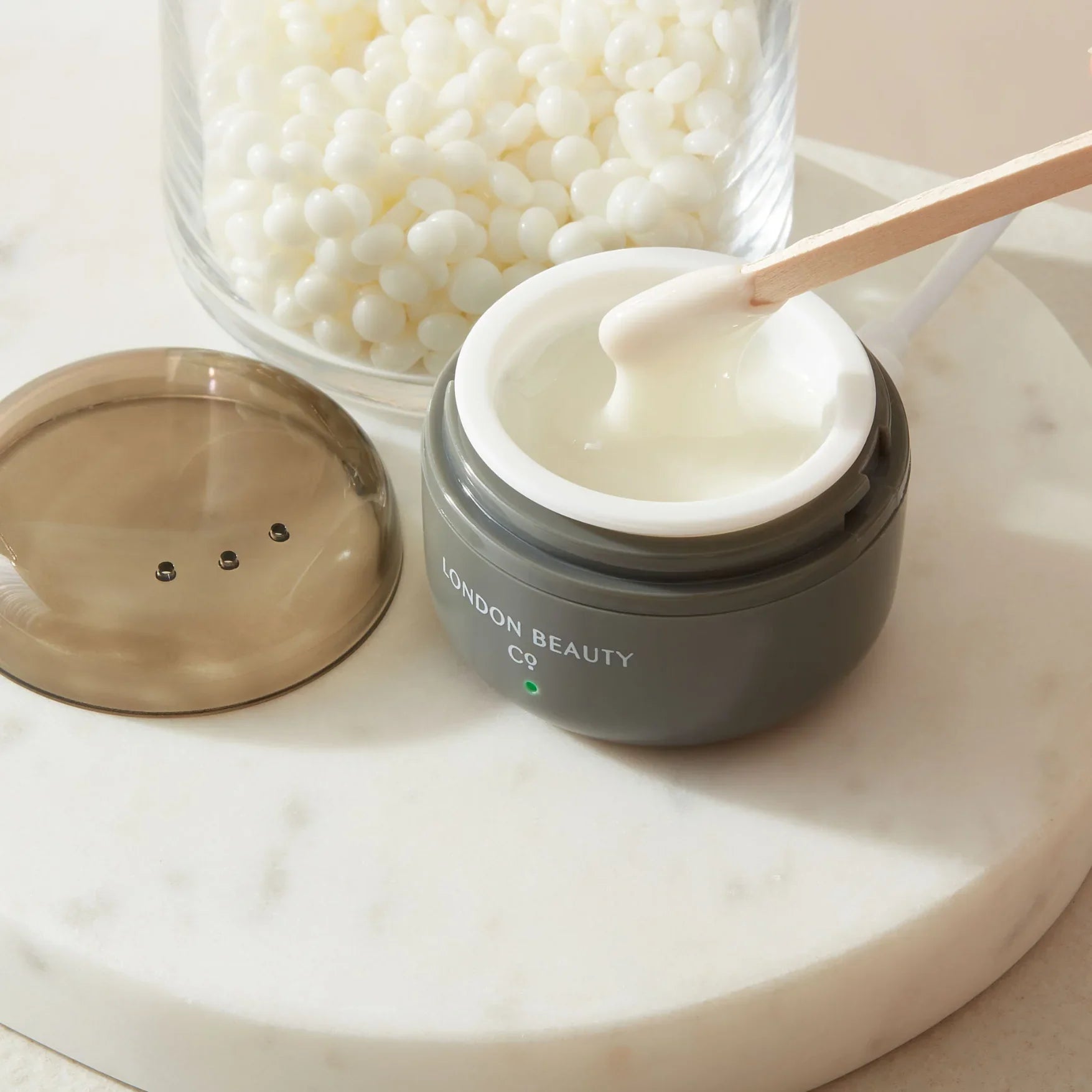 Wax jar with cream, wooden applicator, and pearls on a marble surface