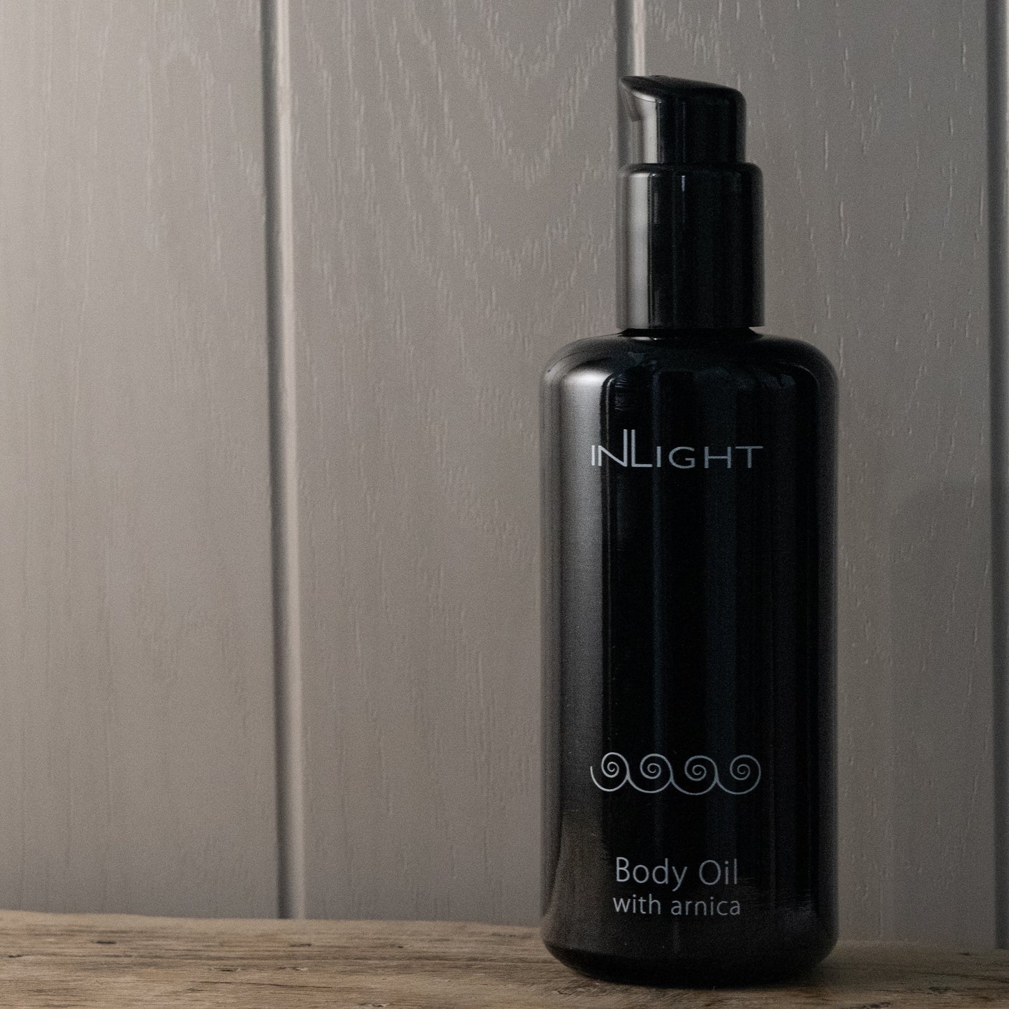 Black bottle of INLIGHT Body Oil with Arnica on a wooden surface with a neutral background