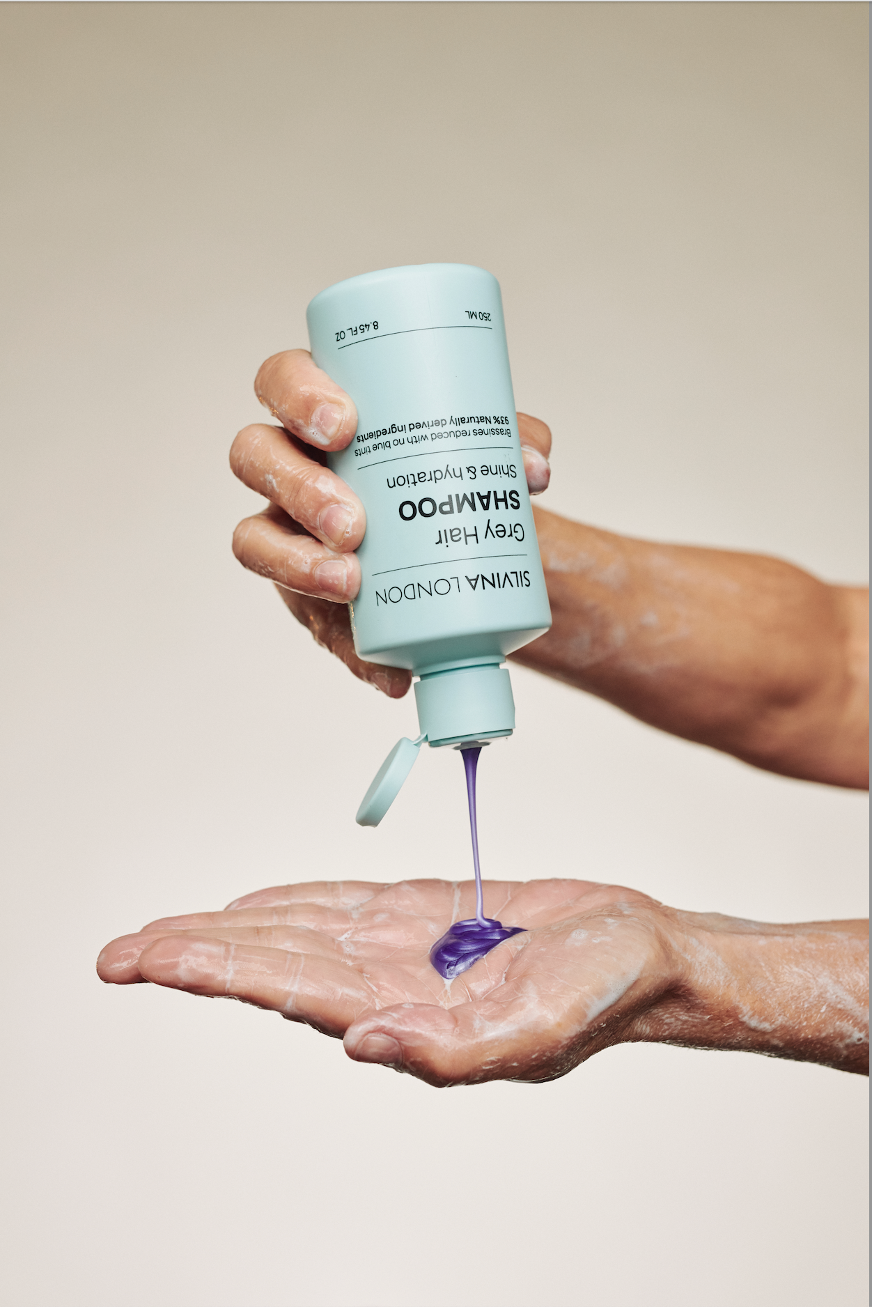 Hand holding a blue bottle of shampoo with purple shampoo being poured onto a palm against a beige background