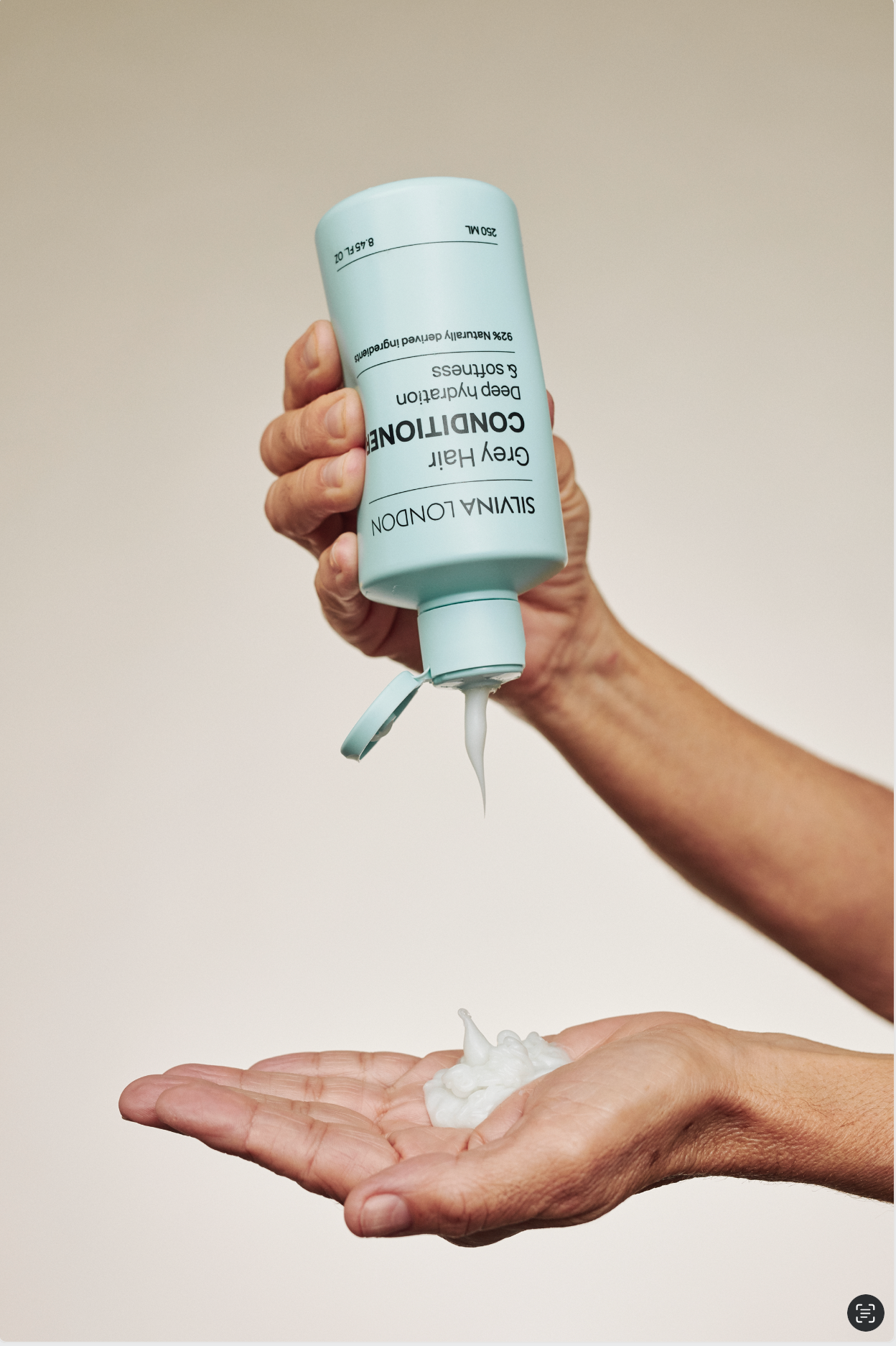Hand holding a bottle of grey hair conditioner with a beige background