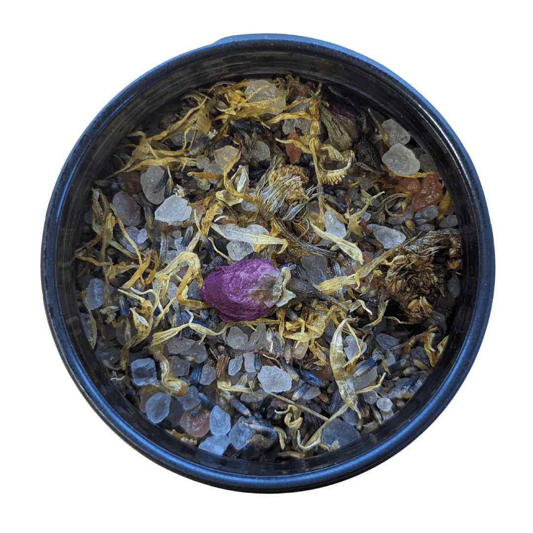 A top-down view of a dark blue ceramic bowl filled with dried botanical tea ingredients, including a prominent purple rosebud, on a solid white background.