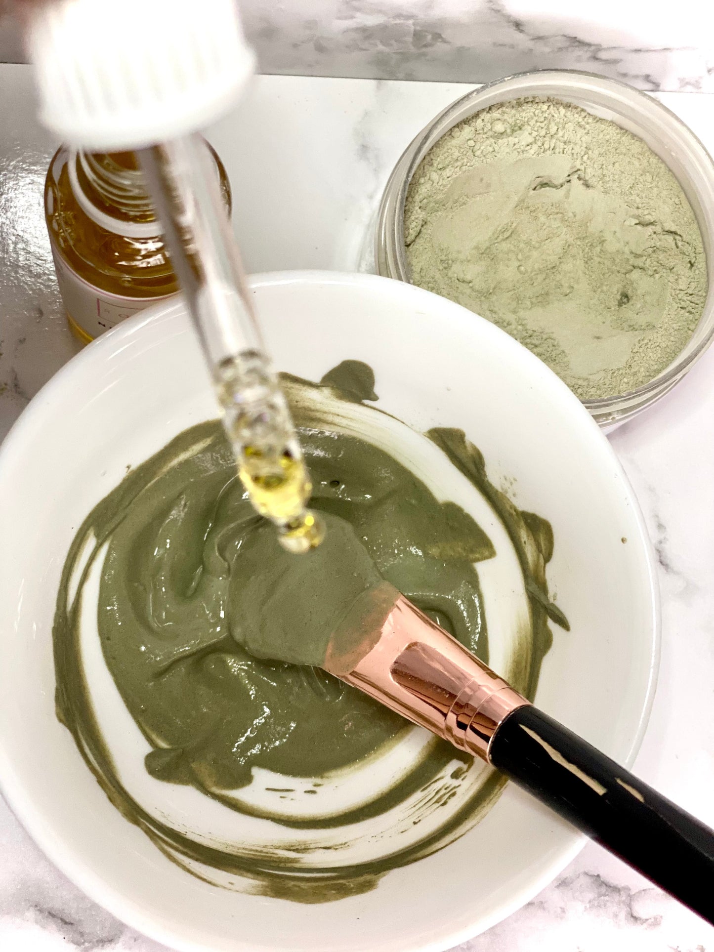 Green facial mask being prepared with a brush and dropper on a marble surface.