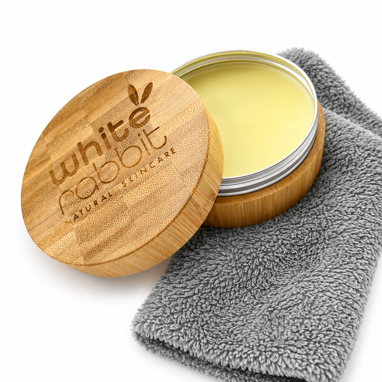 An open round bamboo tin of White Rabbit Natural Skincare balm with pale yellow cream visible inside, shown next to a folded grey microfiber cloth on a white background.