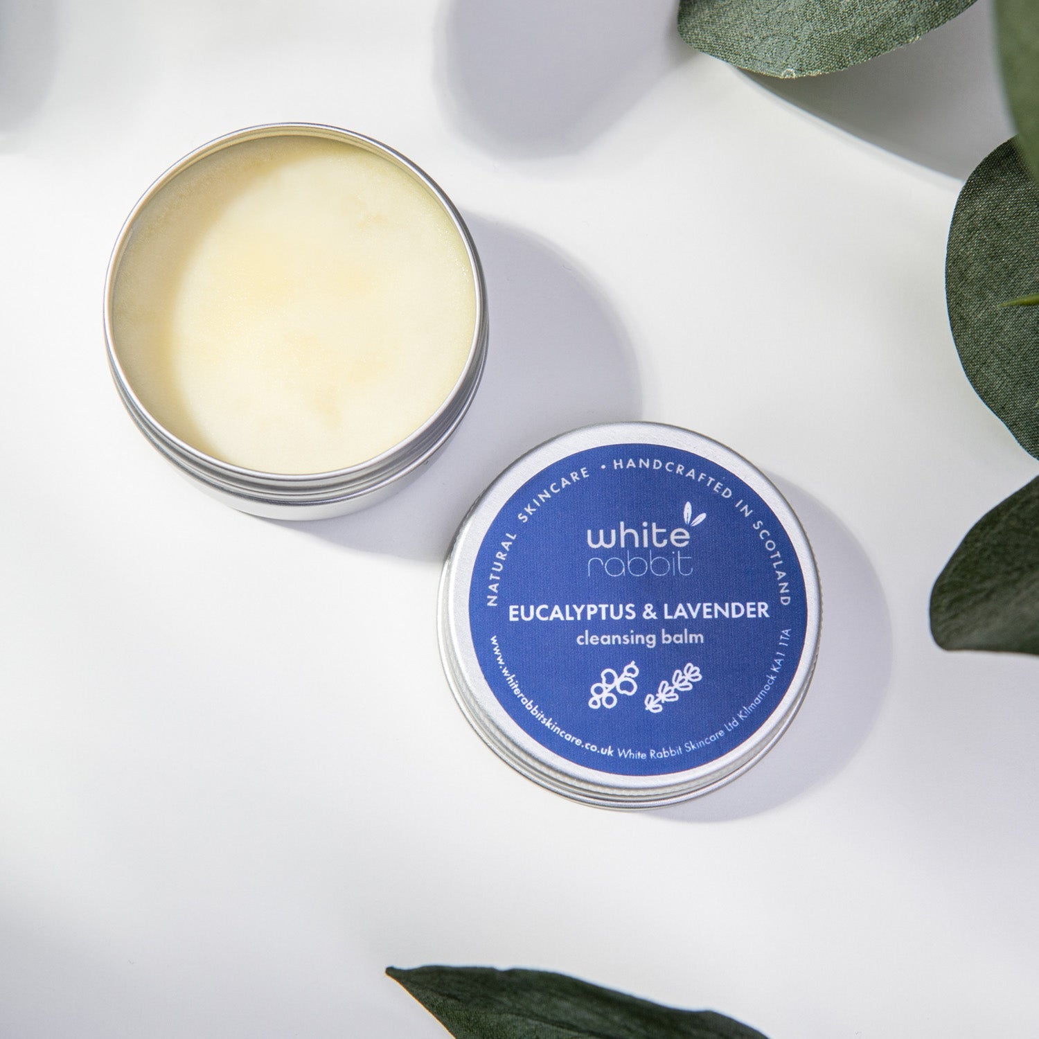 Open tin of White Rabbit Eucalyptus & Lavender cleansing balm on a white background with green leaves.