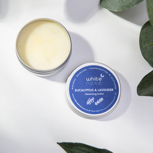 Open tin of White Rabbit Eucalyptus & Lavender cleansing balm on a white background with green leaves.