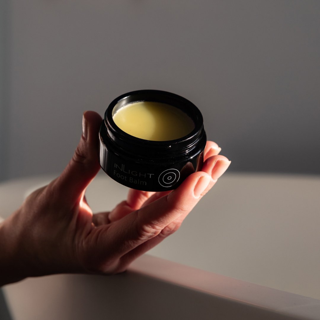 Hand holding a black jar of Inlight Beauty Foot Balm 45ml against a neutral background