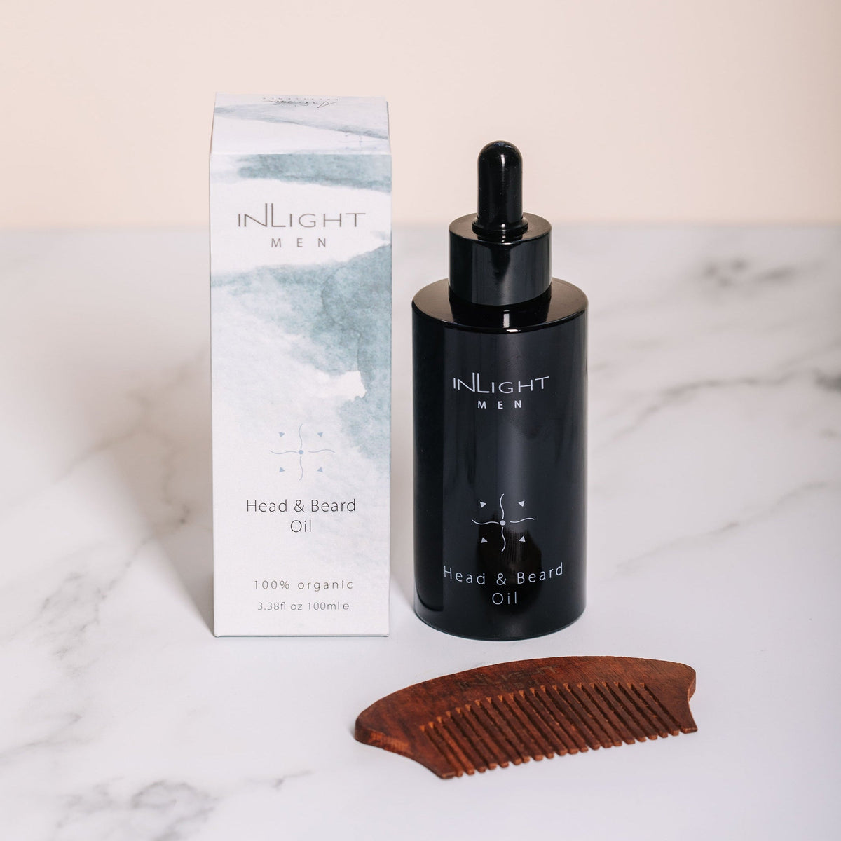 Inlight Beauty Head & Beard Oil for Men 100ml in a black dropper bottle next to its white and blue-green box, with a wooden comb in front, set on a white marble surface.