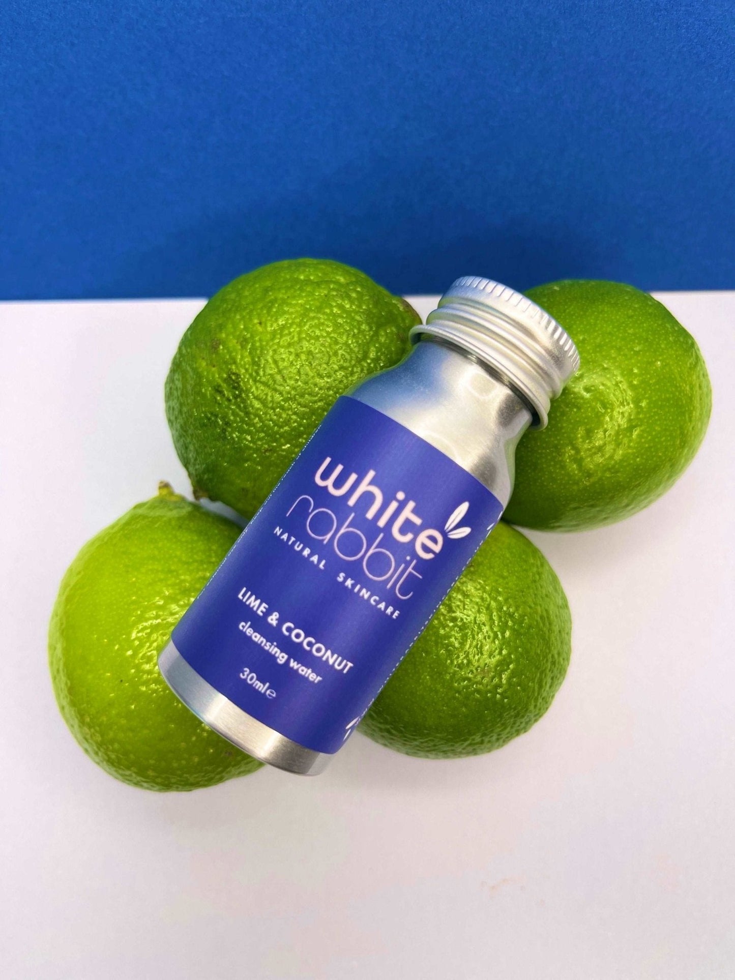 White Rabbit skincare product bottle with lime fruits on a white surface against a blue background
