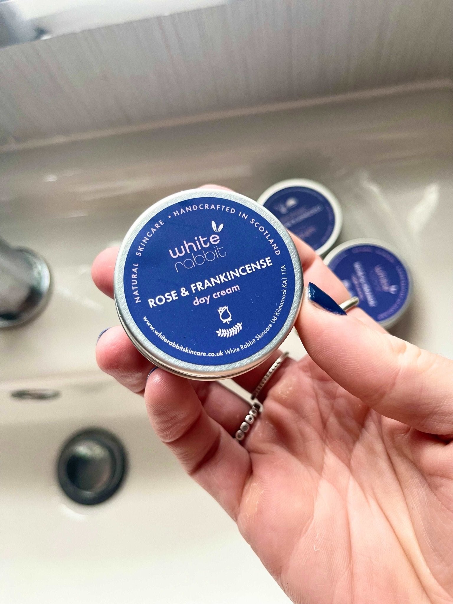 Hand holding a tin of White Rabbit Rose & Frankincense day cream over a sink.