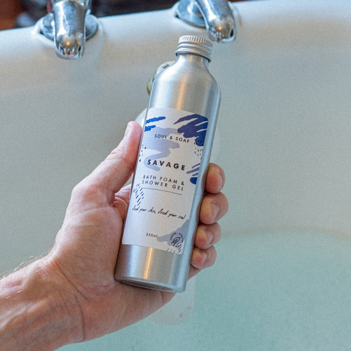Hand holding a metallic bottle labeled 'Savage' in front of a sink.