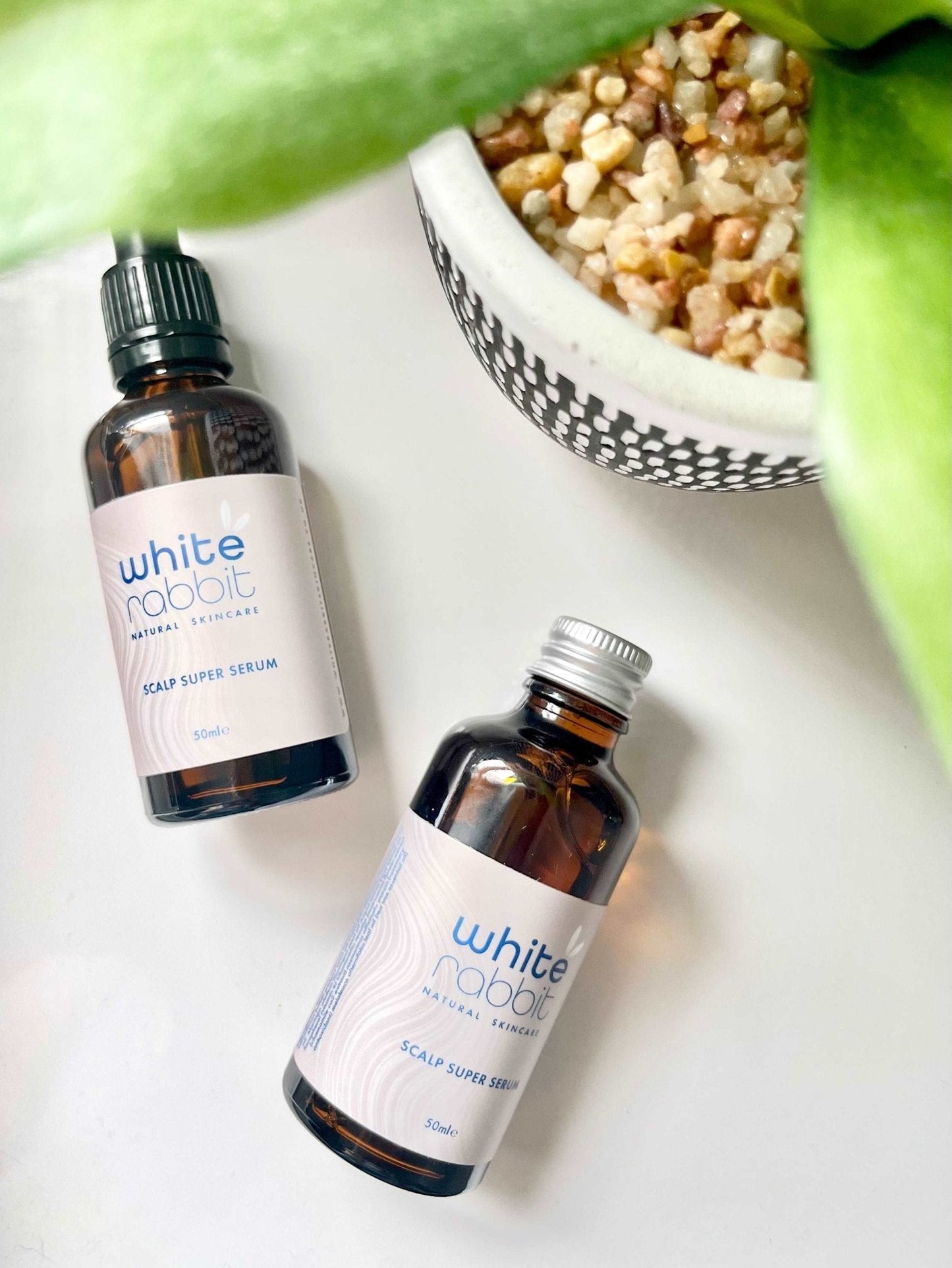 Two bottles of 'White Rabbit' scalp serum on a white surface with a bowl of cereal and green leaves in the background.