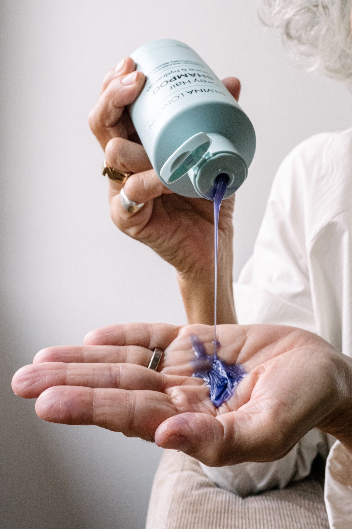 Person pouring grey hair purple shampoo from a bottle into their palm against a neutral background