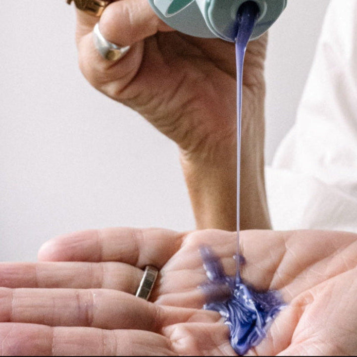 Hand holding a small container with purple liquid being poured onto another hand.