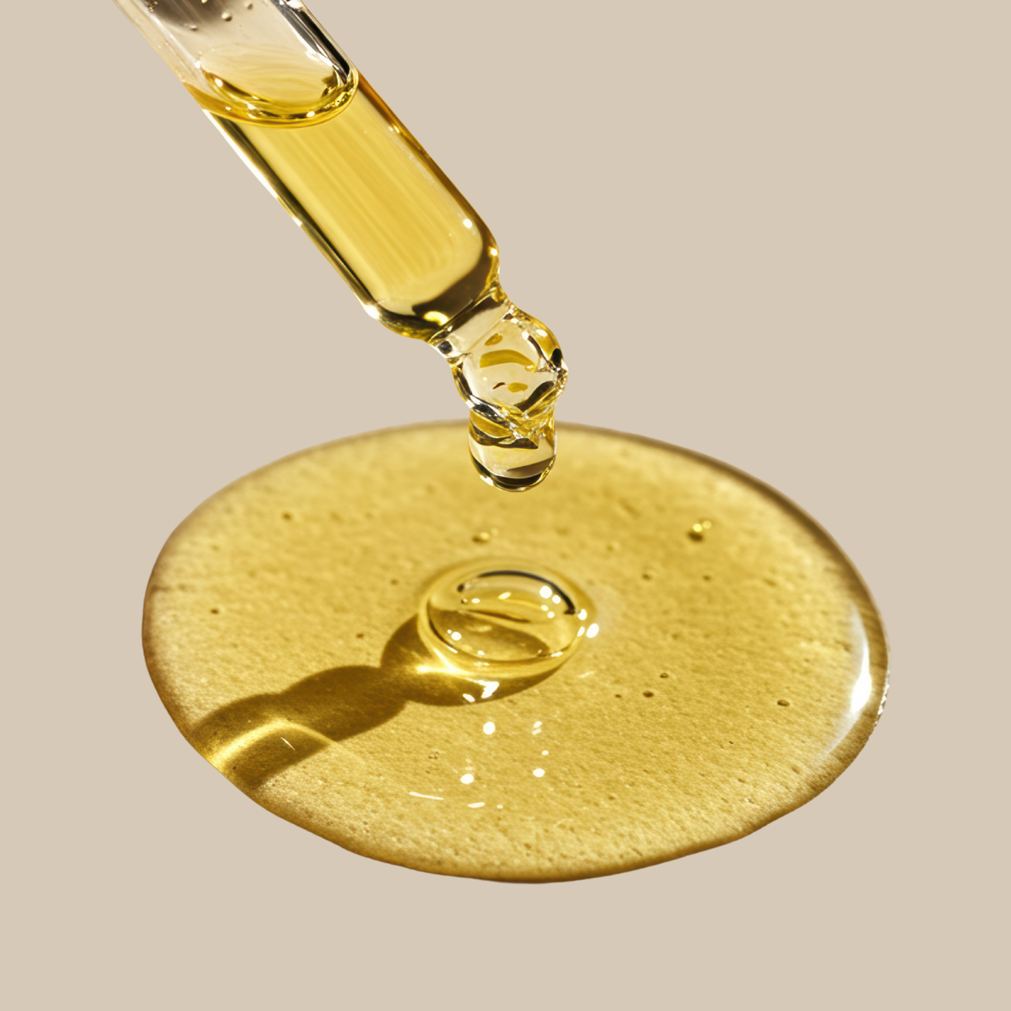 A close-up of a glass dropper releasing a glossy golden-yellow liquid that pools in a circular puddle on a warm beige background.