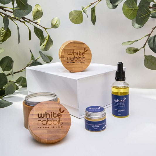 White Rabbit natural skincare products including a candle, jar, and bottles on a white surface with green leaves in the background.
